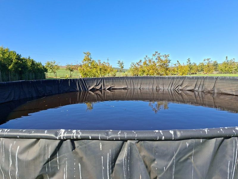 A large circular agricultural water storage tank with a black liner, situated in an open farm landscape.