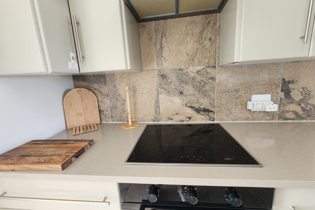 A modern kitchen workspace featuring a built-in electric hob, oven, extractor fan, and stone-look tiled splashback with neutral cabinetry.
