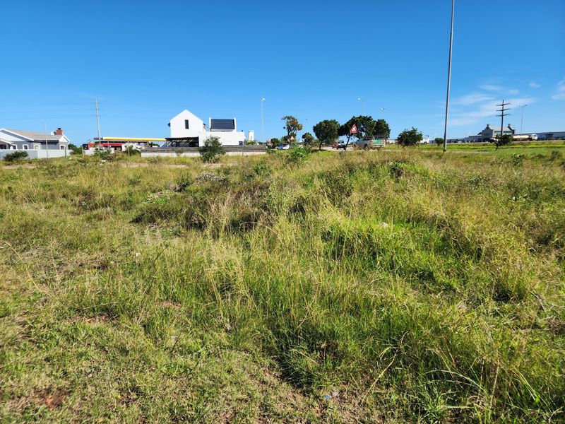 A view of a vacant residential plot featuring natural grass coverage, with surrounding neighborhood infrastructure and nearby buildings visible in the background.