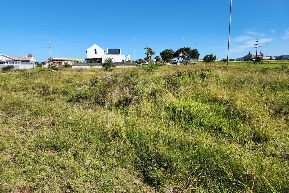 A view of a vacant residential plot featuring natural grass coverage, with surrounding neighborhood infrastructure and nearby buildings visible in the background.