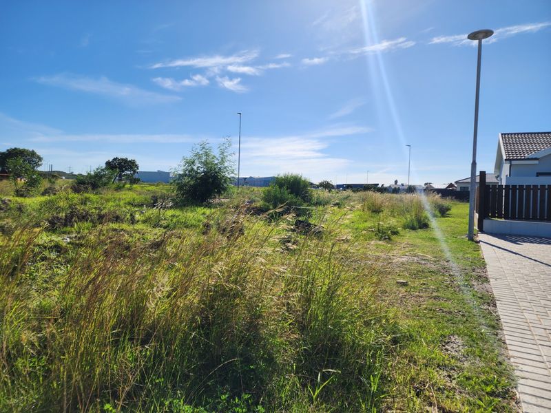 A vacant residential plot featuring natural grass, shrubs, and a paved walkway adjacent to an existing property boundary.