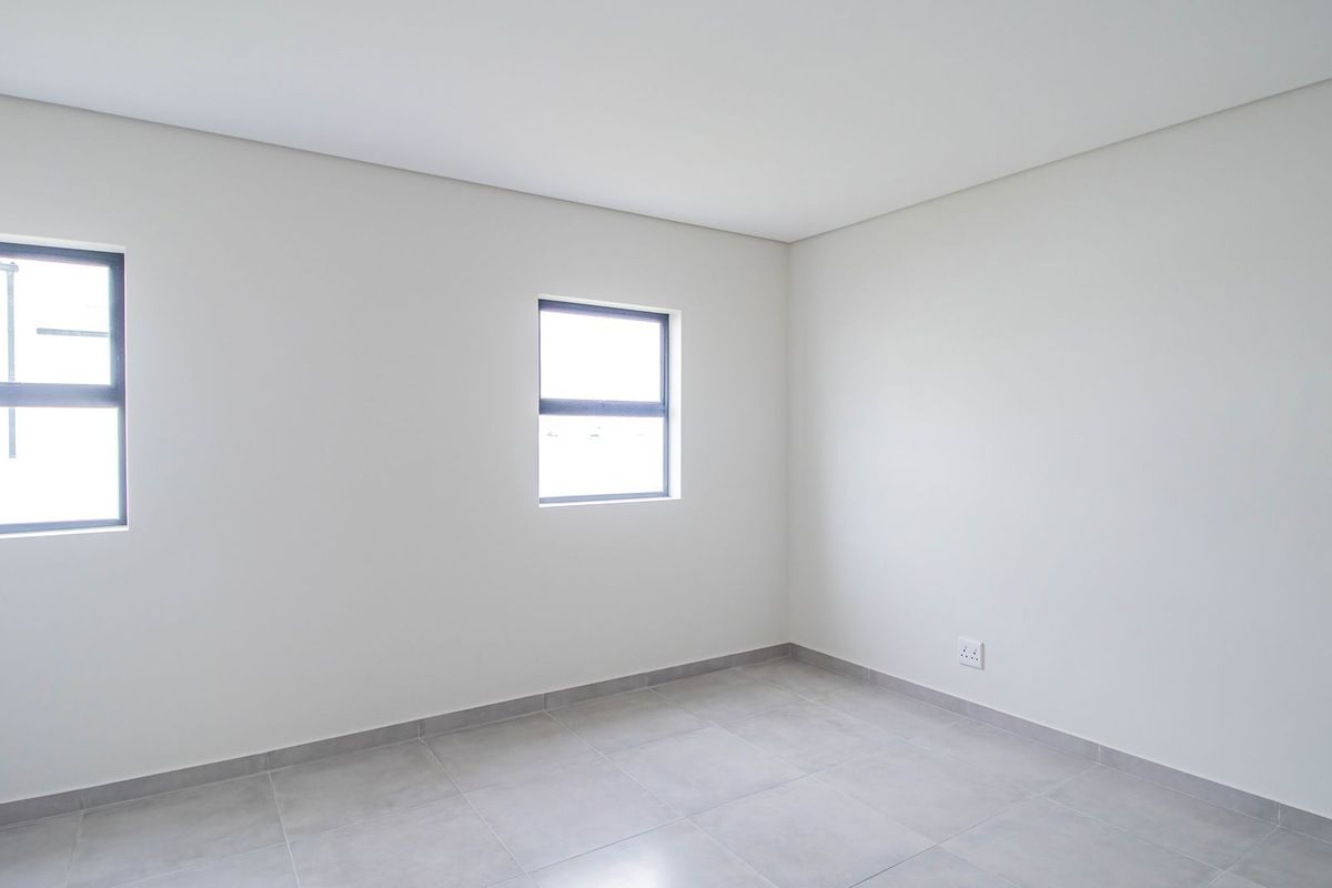 A bright, empty bedroom featuring neutral wall finishes, tiled flooring, and modern aluminium-framed windows.