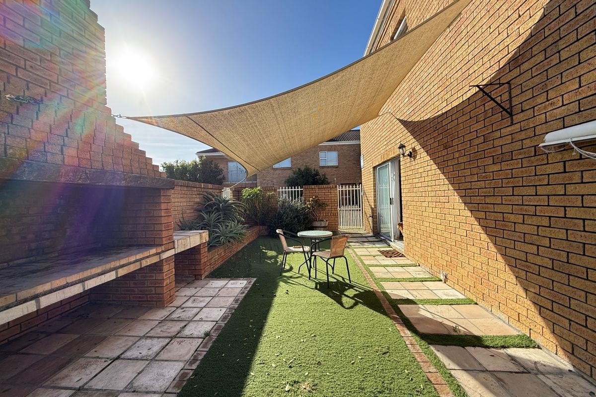 A rear courtyard featuring a built-in brick braai, artificial lawn, paved pathways, and a shade sail for outdoor entertaining.