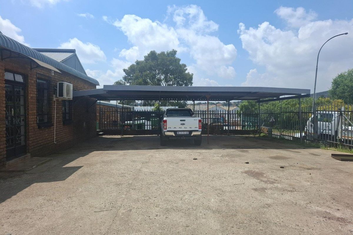 Rear exterior area featuring a large steel carport, concrete paving, and secure perimeter fencing, suitable for industrial parking or utility access.