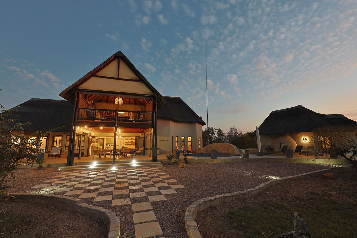 A spacious thatched-roof residence featuring a covered patio, outdoor entertainment area, and a swimming pool set within a landscaped garden at dusk.