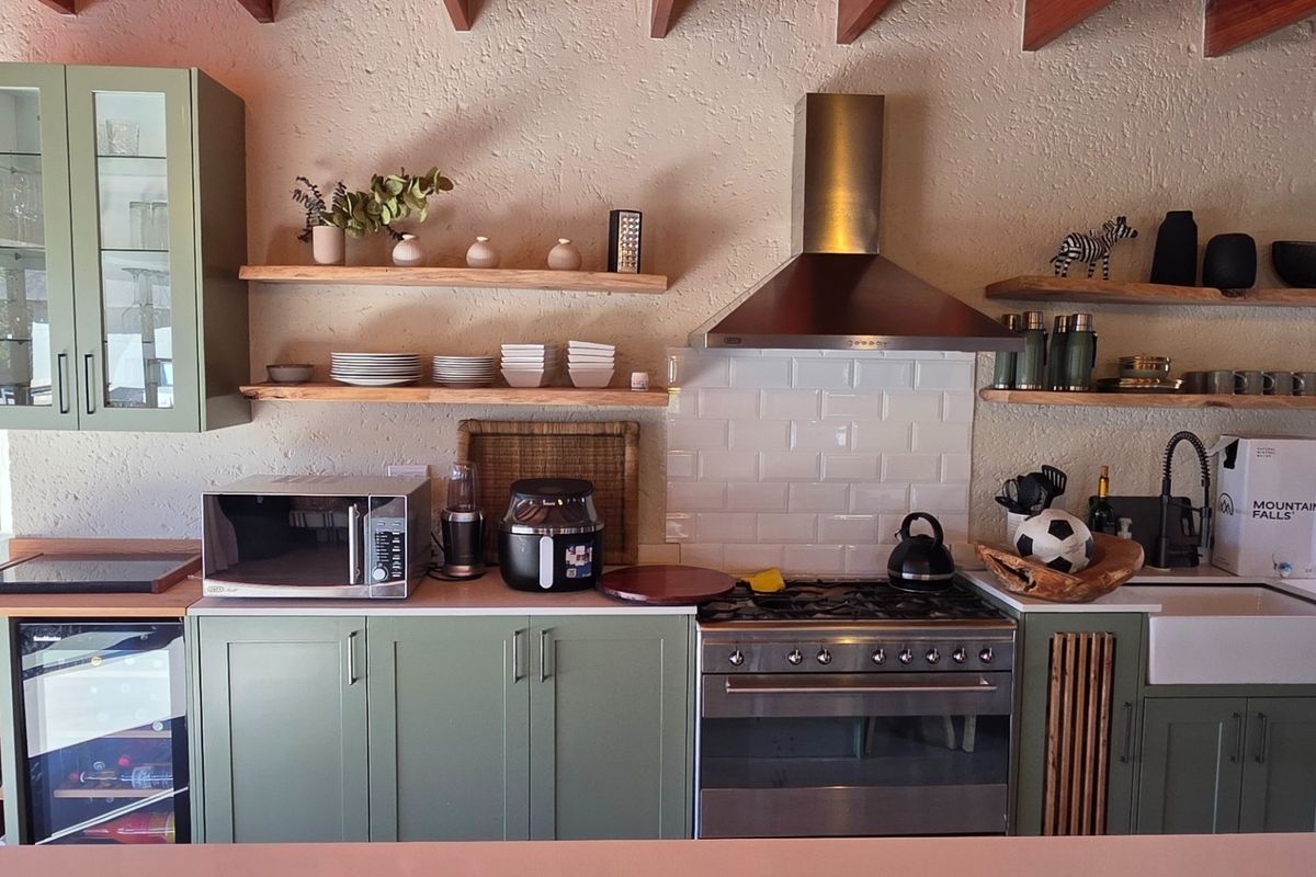 Modern kitchen featuring sage green cabinetry, subway tile backsplash, stainless steel extractor fan, gas stove, and a farmhouse-style sink with exposed timber shelving.