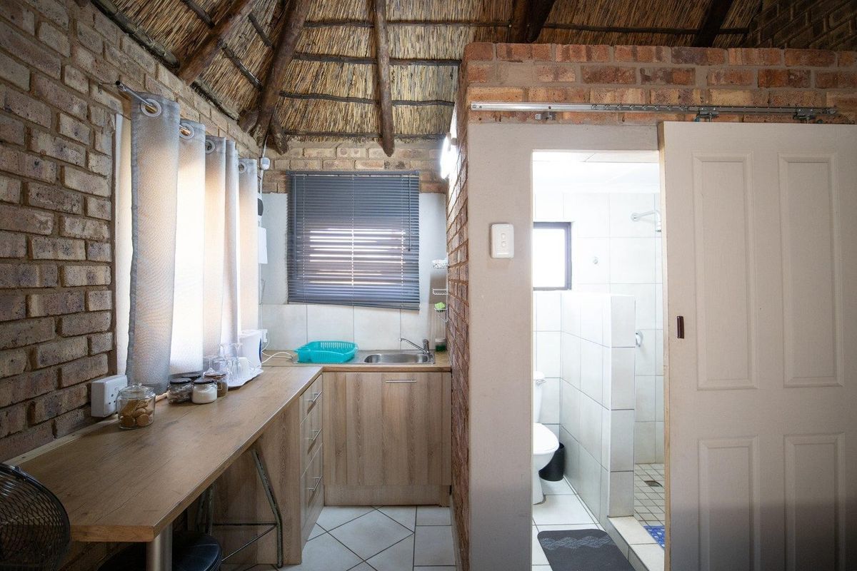 Compact kitchenette area featuring wooden cabinetry and a stainless steel sink, adjacent to a tiled bathroom with a shower enclosure under a thatched roof.