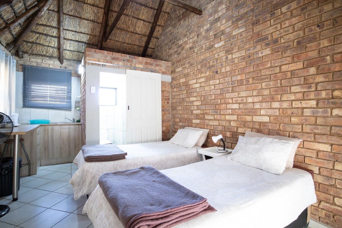 A rustic bedroom featuring exposed brick walls, a high thatched roof, and an integrated kitchenette area with a small en-suite bathroom.