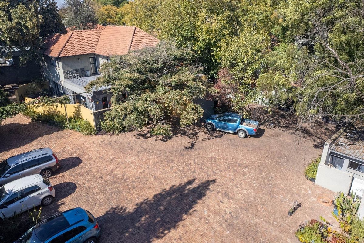 Aerial view of a commercial property featuring a large paved parking area, mature trees, and a multi-level building with a tiled roof.