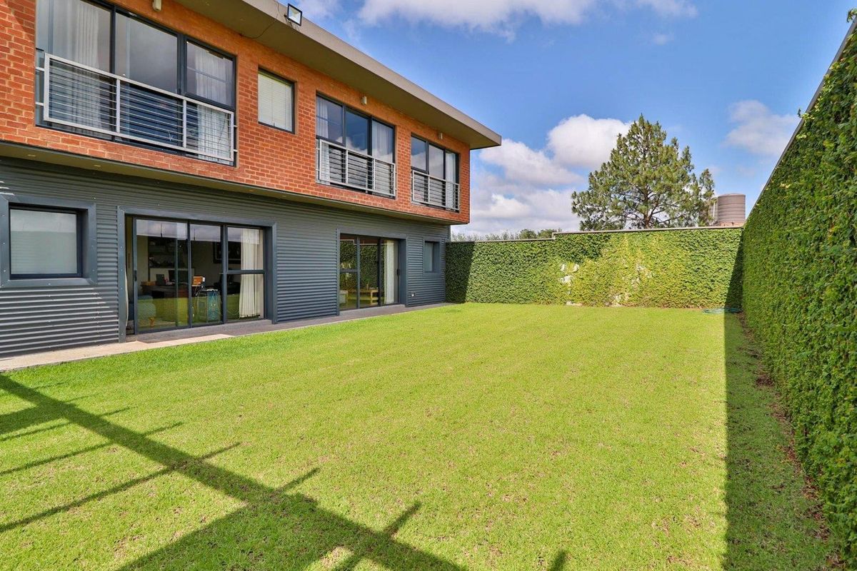 A well-maintained rear garden featuring a lush green lawn, enclosed by a high ivy-covered boundary wall, adjacent to a modern two-story brick and metal-clad home.