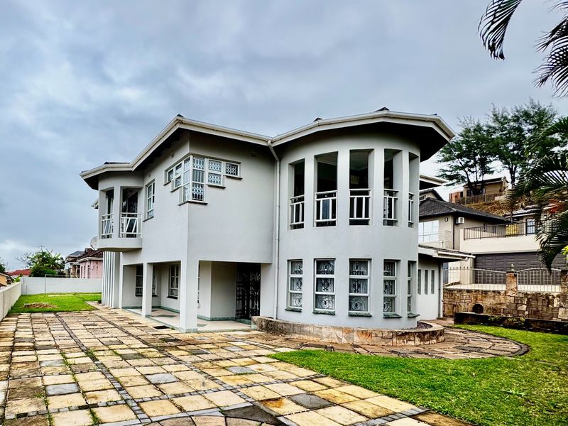 A multi-story residential property featuring a curved architectural design, balcony, and paved driveway area set within a landscaped garden.