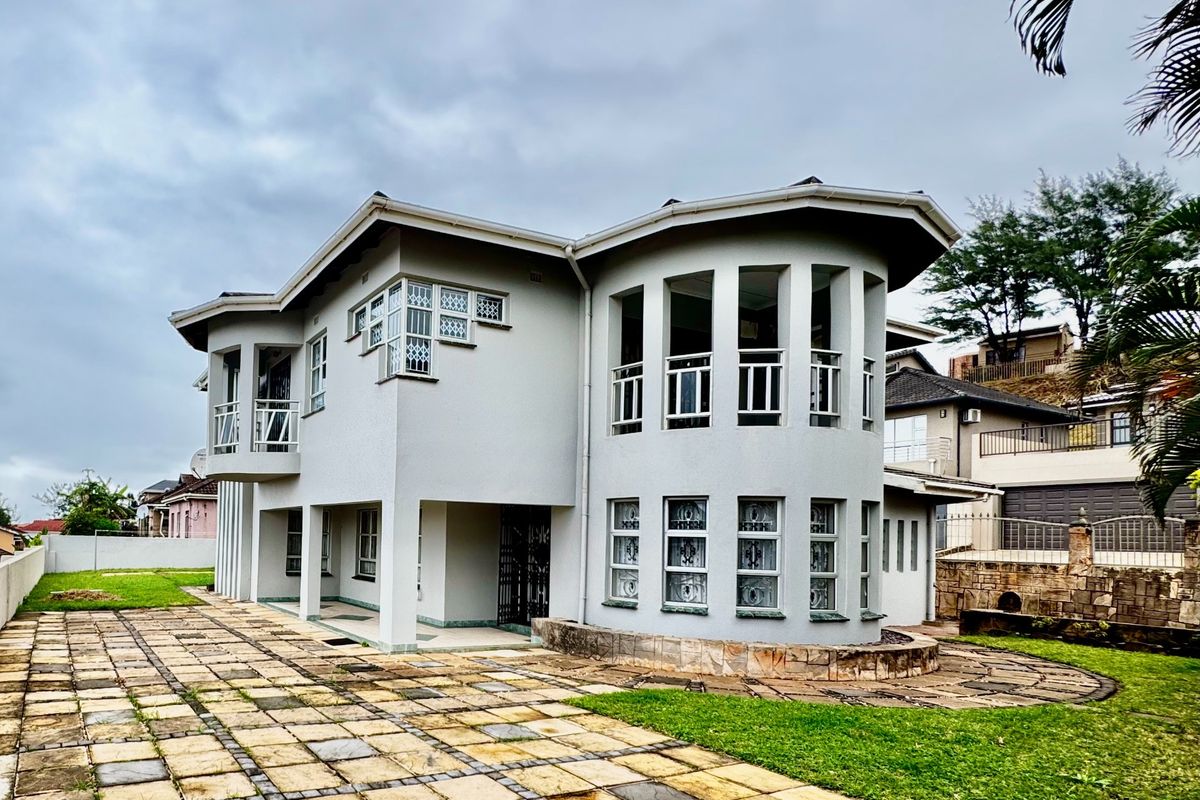 A multi-story residential property featuring a curved architectural design, balcony, and paved driveway area set within a landscaped garden.