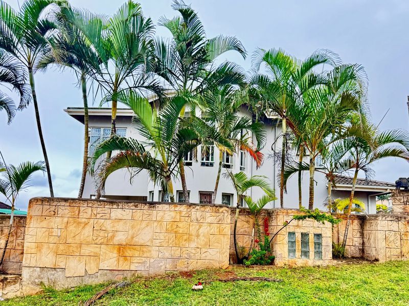 Exterior view of a multi-story house featuring a textured stone boundary wall, established palm trees, and a landscaped garden area.