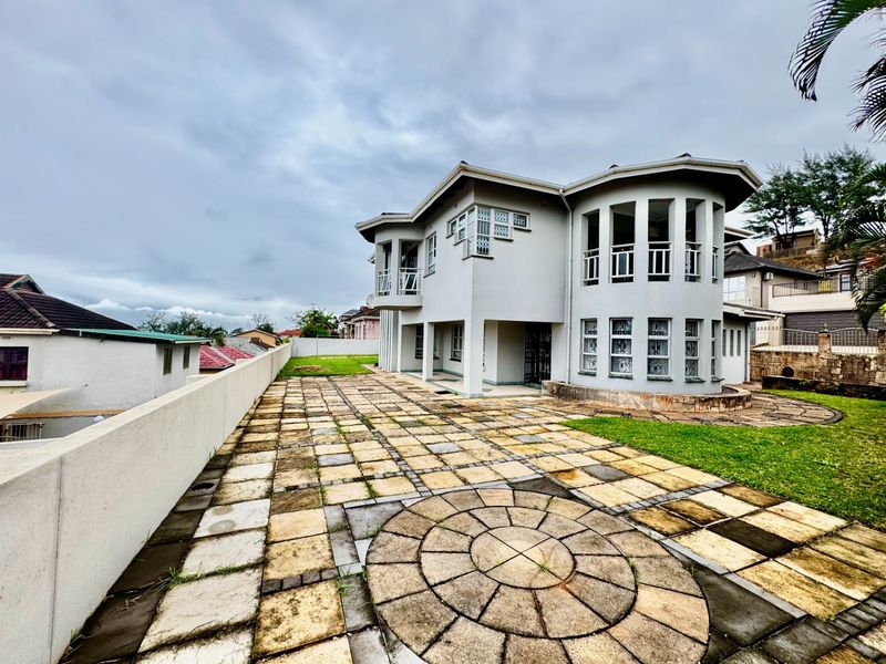 A spacious rear exterior view of a multi-story house featuring a large paved patio area, landscaped lawn, and architectural curved balcony structures.