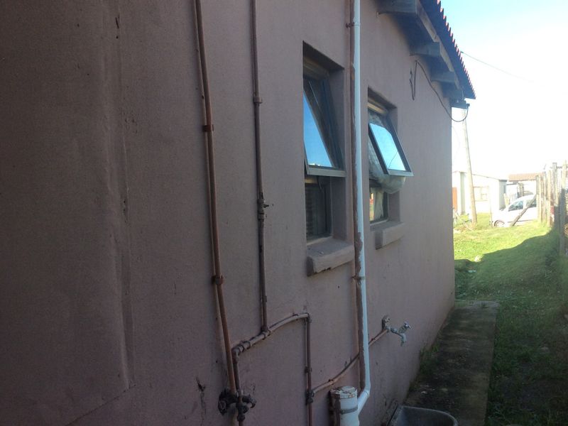 Side view of the property exterior showing exposed plumbing, casement windows, and a concrete pathway along the building perimeter.
