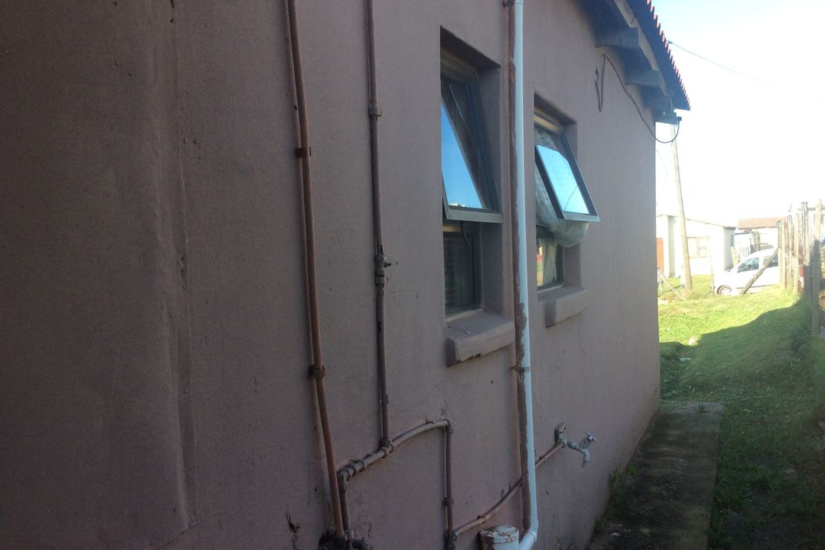 Side view of the property exterior showing exposed plumbing, casement windows, and a concrete pathway along the building perimeter.