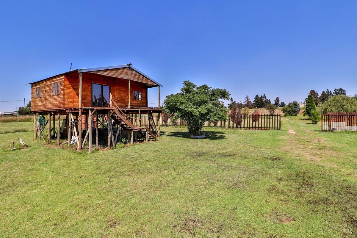 A rustic elevated wooden cabin set on a spacious rural farm plot, featuring a raised deck and expansive surrounding lawn.