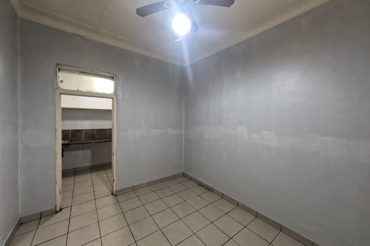 An empty room with tiled flooring and a ceiling fan, featuring an open doorway leading to a secondary kitchen or utility area.