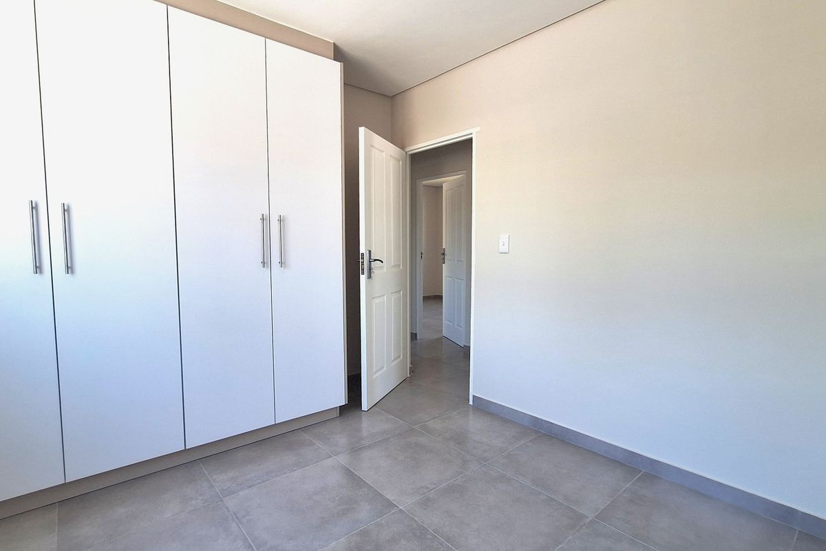 A clean, modern bedroom featuring neutral tiled flooring and built-in white cabinetry with sleek handles, leading into a hallway.