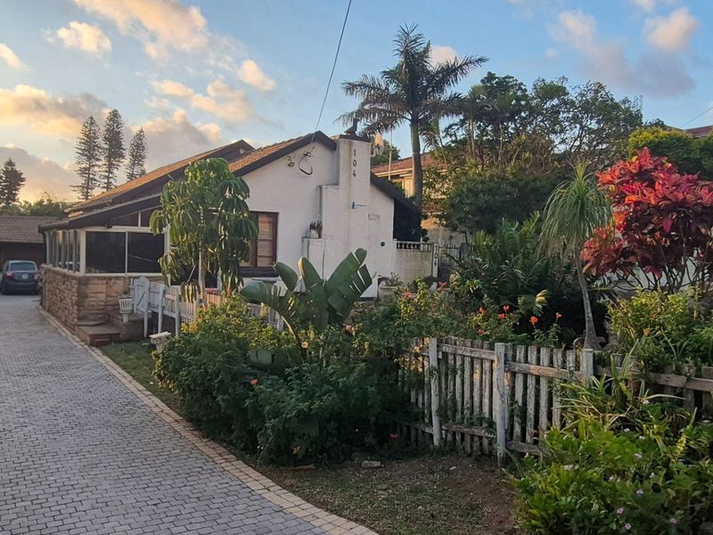 Front view of a residential property featuring a paved driveway, established garden, and a white exterior facade with a chimney.