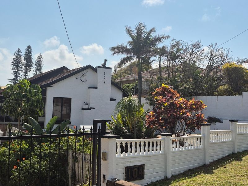 Front view of a white-painted house featuring a secure gated entrance, paved driveway, and a decorative brick boundary wall with balustrade detailing.