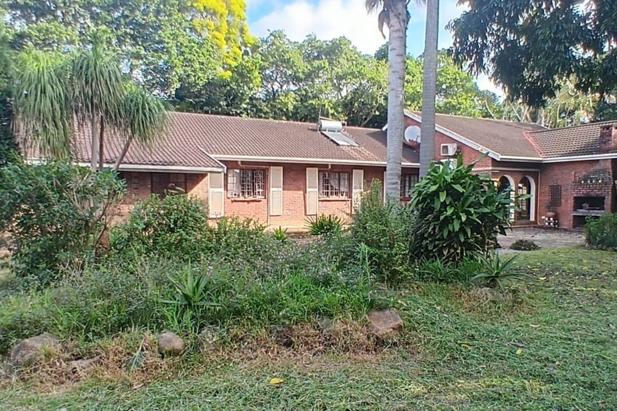 A rear view of a single-story brick house featuring a tiled roof, built-in outdoor braai area, and a spacious, established garden setting.