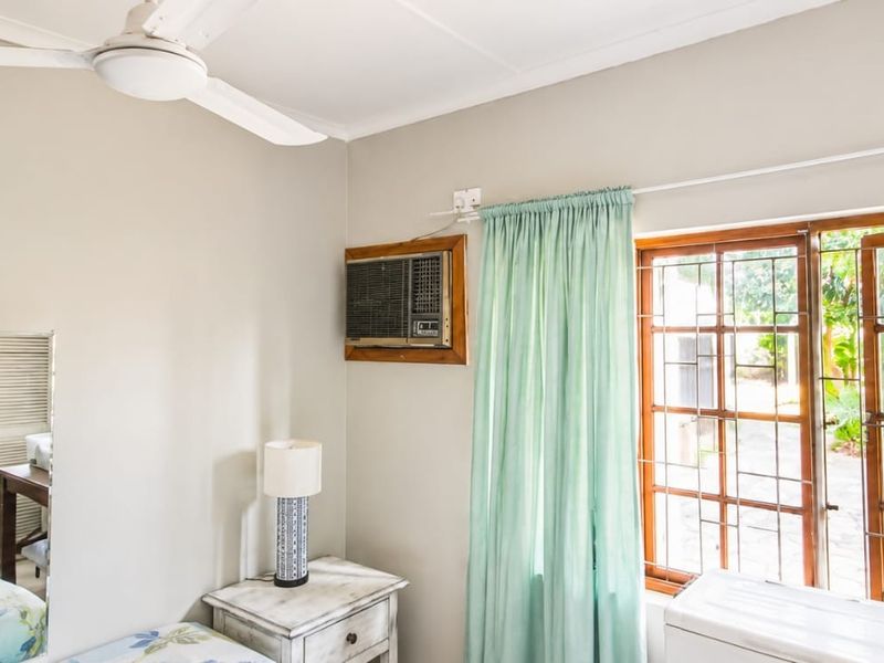 A bedroom featuring a ceiling fan, built-in shelving, and a window air conditioning unit, with natural light entering through wooden-framed windows.