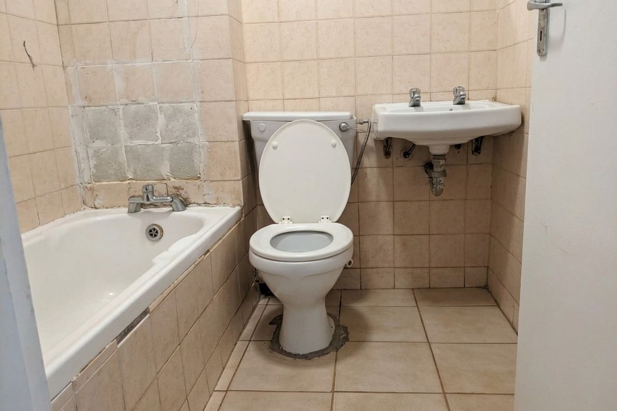 A dated bathroom featuring a standard bathtub, wall-mounted basin, and toilet, requiring significant renovation and tile repairs to modernize the space.