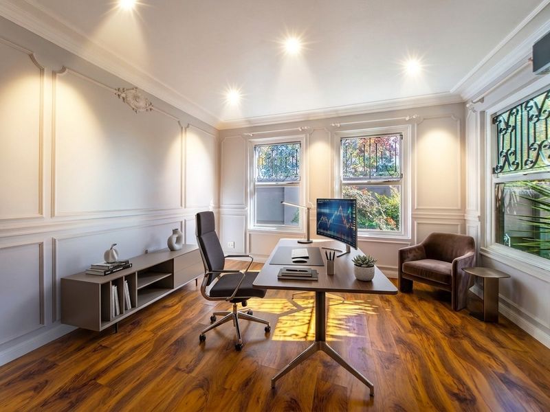 A sophisticated home office featuring elegant wainscoting, rich hardwood flooring, crown molding, and recessed lighting, complemented by large windows with security bars.
