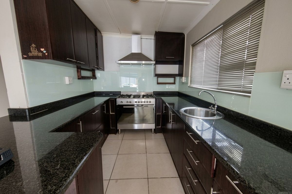 A functional galley-style kitchen featuring dark cabinetry, granite countertops, a glass splashback, and a stainless steel gas stove with an extractor fan.