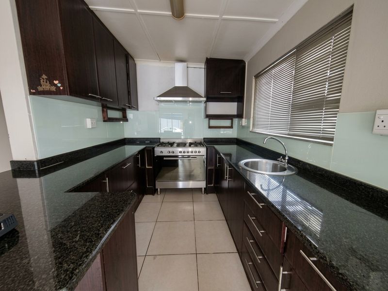 A functional galley-style kitchen featuring dark cabinetry, granite countertops, a glass splashback, and a stainless steel gas stove with an extractor fan.