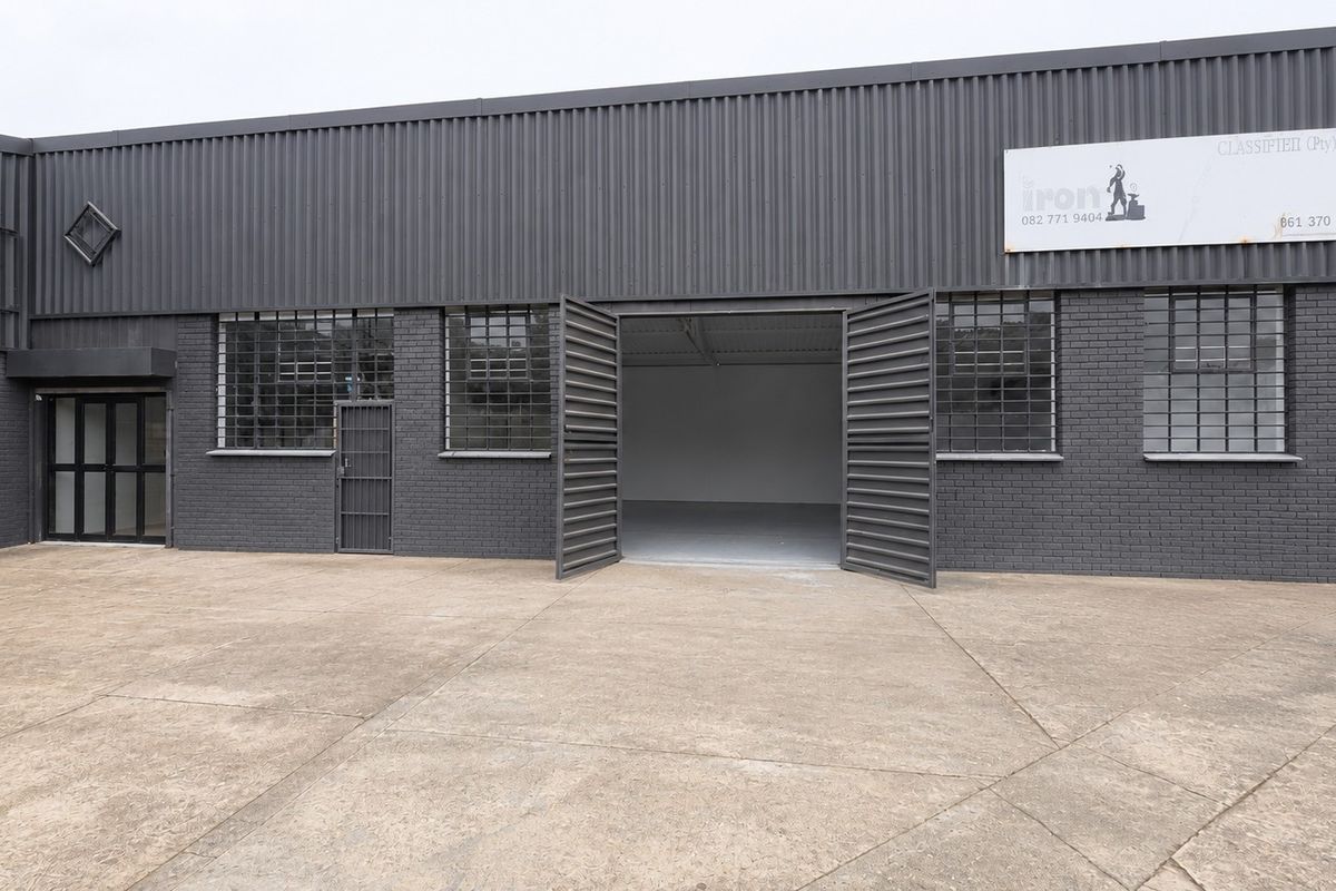 Front elevation of an industrial warehouse featuring dark grey corrugated metal cladding, brickwork, security-barred windows, and large swing doors opening to a concrete yard.