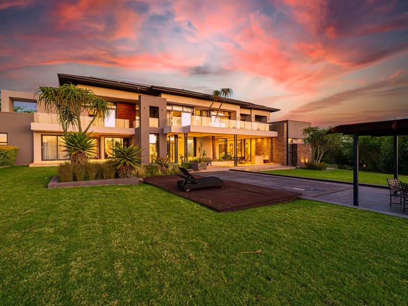 A stunning rear exterior view of a modern two-story luxury residence featuring expansive glass balconies, stone cladding, and a beautifully landscaped garden.