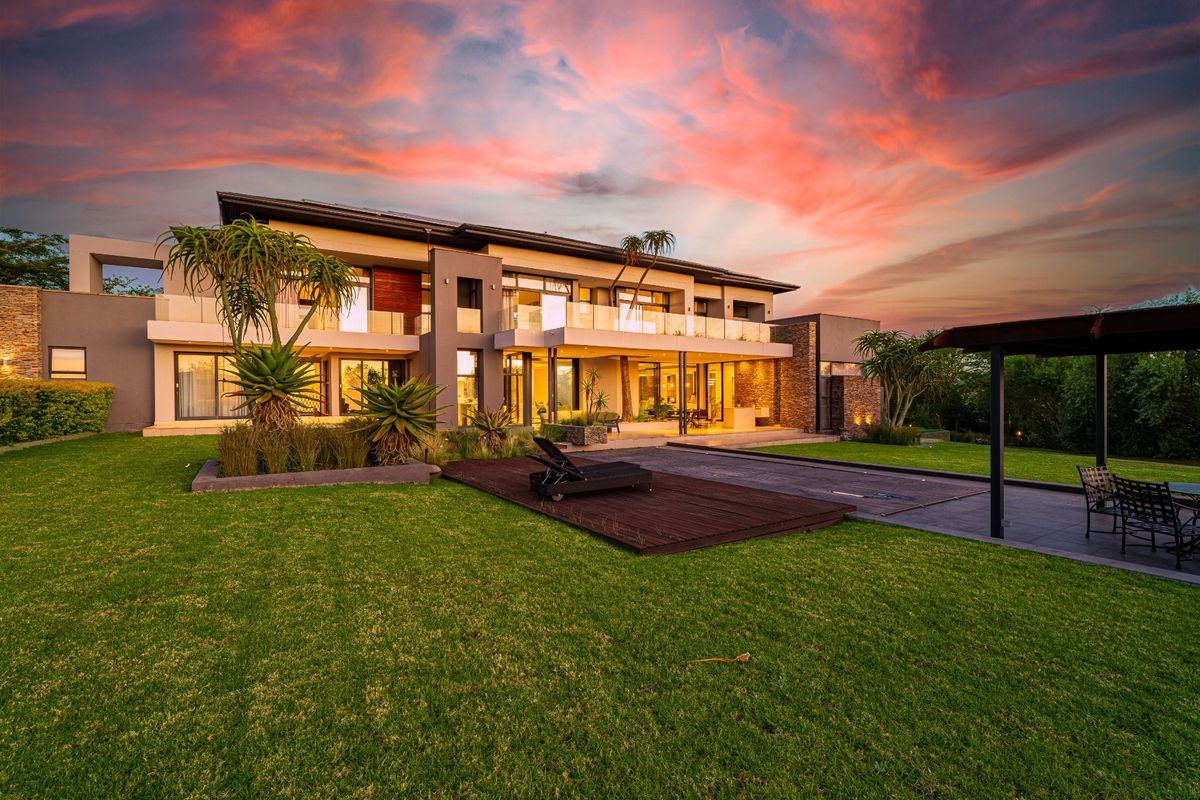 A stunning rear exterior view of a modern two-story luxury residence featuring expansive glass balconies, stone cladding, and a beautifully landscaped garden.