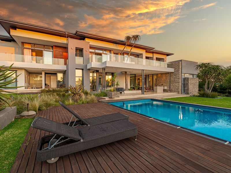 A stunning rear exterior view of a modern luxury home featuring a swimming pool, timber decking, and expansive glass balconies at sunset.
