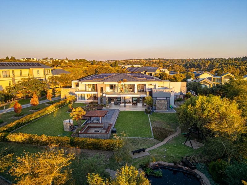 Aerial view of a large modern residence featuring a landscaped garden, sunken fire pit area, covered patio, and solar panel installation on the roof.