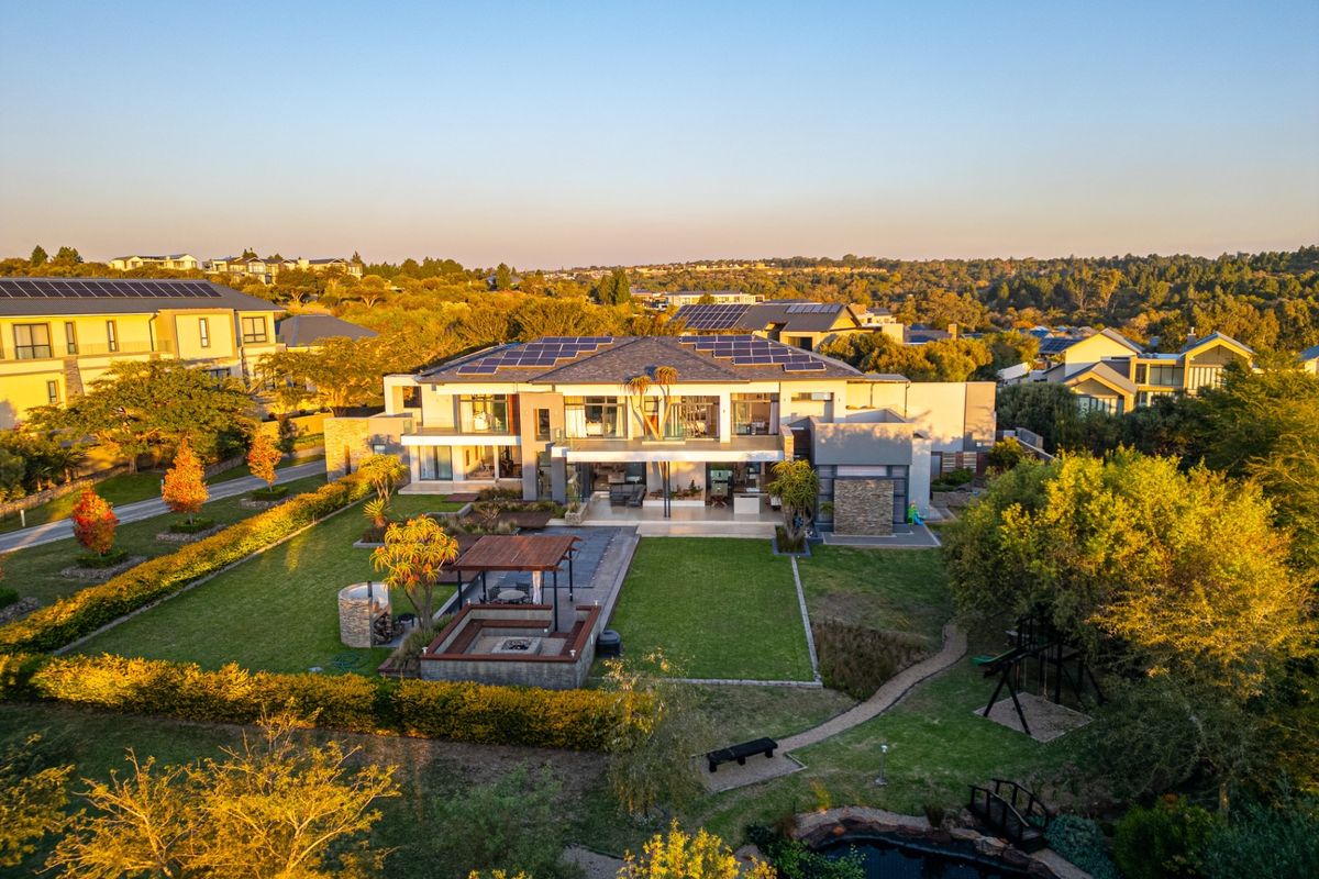 Aerial view of a large modern residence featuring a landscaped garden, sunken fire pit area, covered patio, and solar panel installation on the roof.