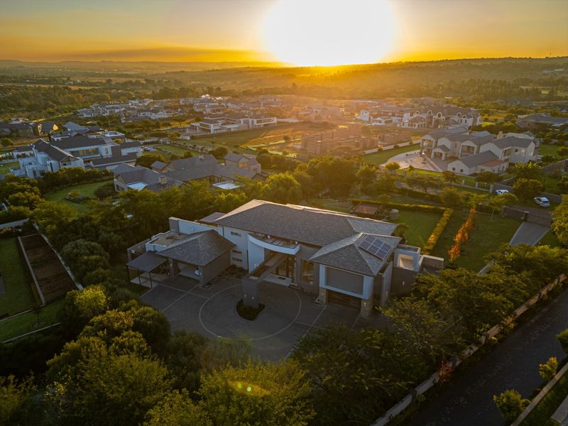 Aerial view of a modern residential property featuring a paved circular driveway, multi-car garage, and solar panels, set within a landscaped estate at sunset.