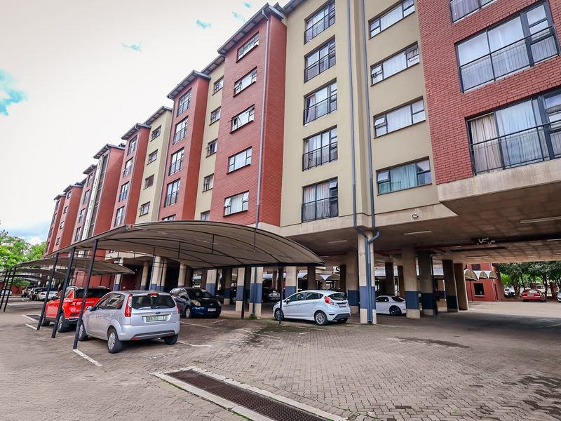 Exterior view of a multi-story apartment complex featuring a paved parking area with covered bays and concrete structural supports.