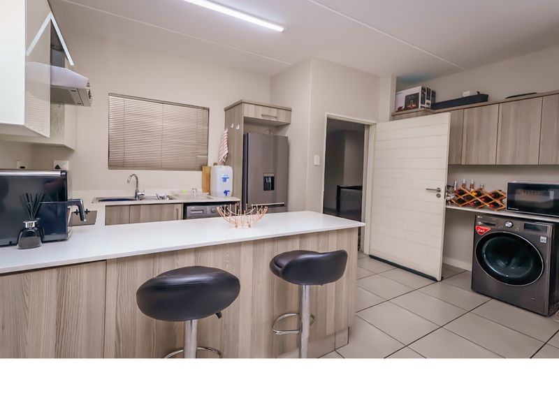 Modern open-plan kitchen featuring light wood-grain cabinetry, stone countertops, integrated laundry space, and tiled flooring in a well-maintained apartment setting.