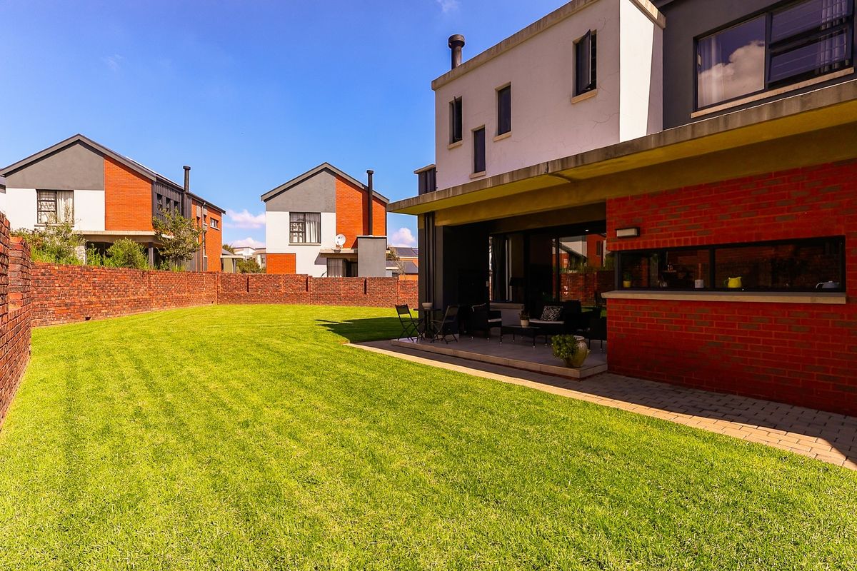 A spacious rear garden featuring a well-maintained lawn, a paved patio area, and a modern house with exposed brick and rendered finishes.