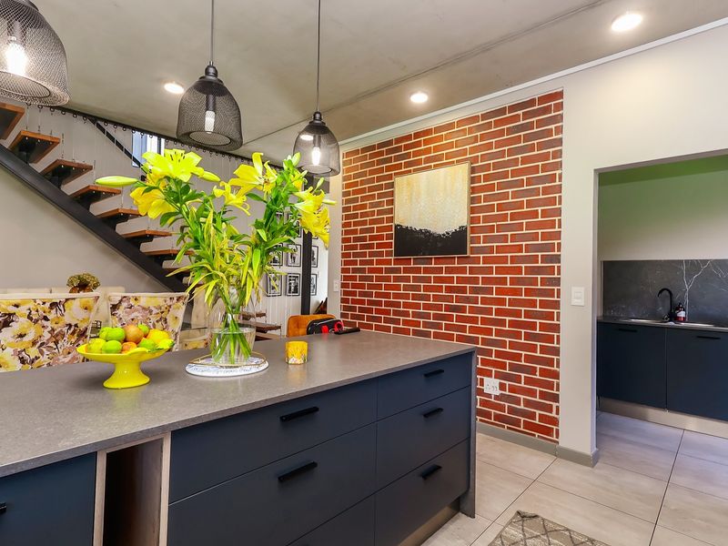 Modern open-plan kitchen featuring a central island, exposed brick feature wall, and an adjacent scullery, complemented by contemporary pendant lighting and a staircase.