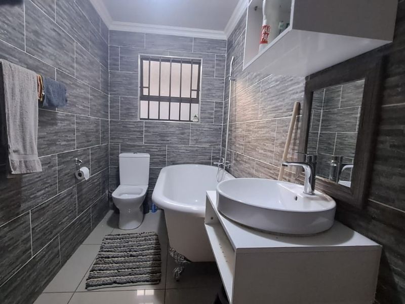 A modern bathroom featuring grey textured wall tiles, a freestanding clawfoot bathtub, a white vanity with a vessel sink, and a security-barred window.
