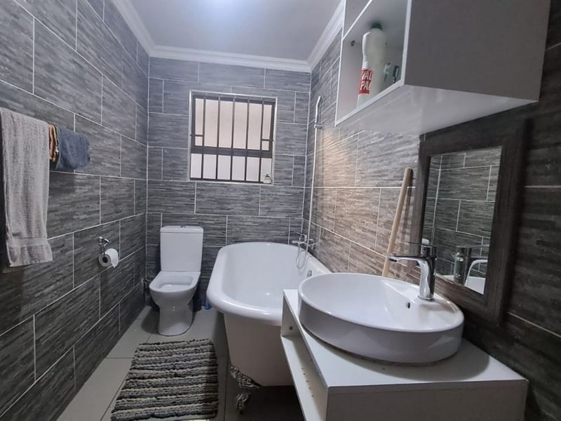 A modern bathroom featuring grey textured wall tiles, a freestanding clawfoot bathtub, a white vessel sink on a vanity, and a toilet.