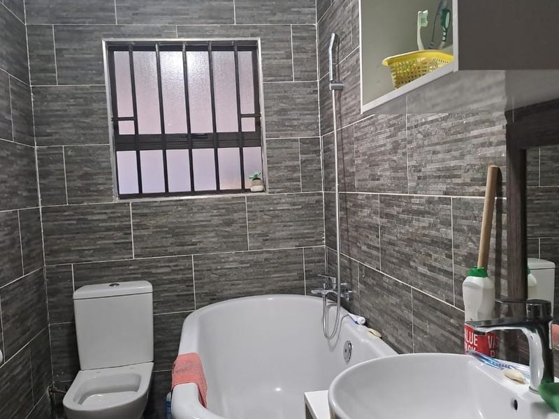 A compact bathroom featuring full-height grey stone-look wall tiles, a built-in bathtub with handheld shower, a modern basin, and a toilet.