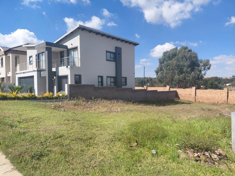 A view of a modern residential property featuring a multi-story home with a balcony, adjacent to an undeveloped grassy plot with a brick boundary wall.