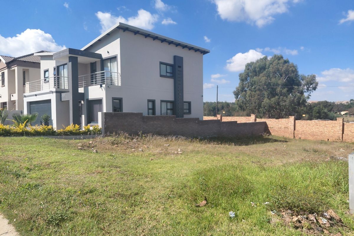 A view of a modern residential property featuring a multi-story home with a balcony, adjacent to an undeveloped grassy plot with a brick boundary wall.