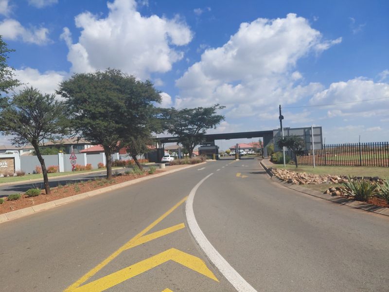 View of the secure estate entrance gate and access road, featuring paved surfaces, perimeter fencing, and established landscaping.