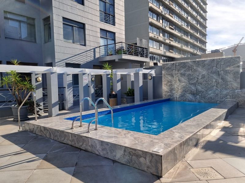 A well-maintained outdoor pool area featuring tiled surrounds, a stone-clad water feature, and a pergola structure adjacent to the apartment building.