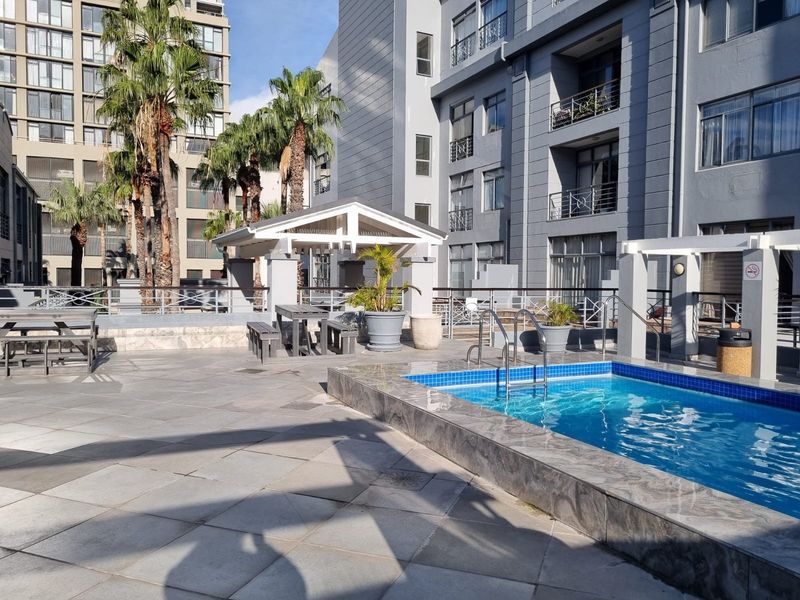 Communal outdoor pool area featuring paved decking, a covered pergola, and modern apartment building architecture.